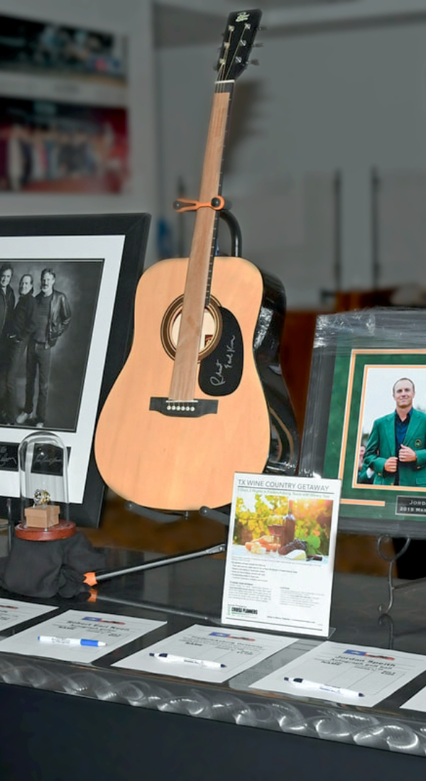 A display table features a signed acoustic guitar, framed photos, a wine country getaway flyer, and several pens and bid sheets, inviting guests to support Texas Horse Racing at this exciting auction or fundraiser event.