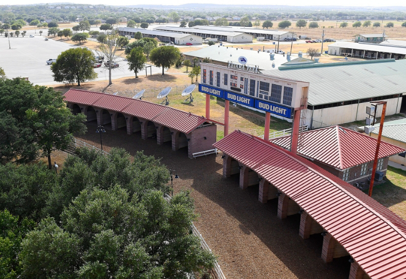 Aerial view of a horse stable with red roofs and outdoor animal pens near the Texas Horse Racing Hall of Fame, featuring a large Bud Light advertising sign, set in a rural area with nearby buildings and parking lot.