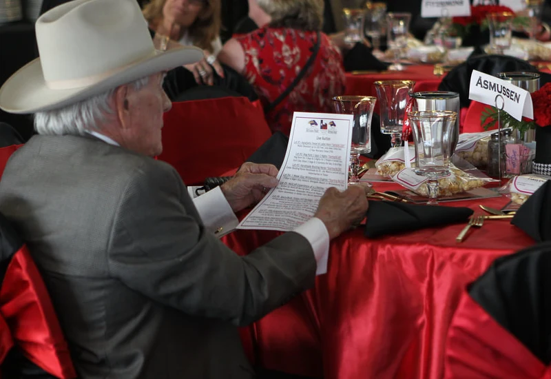 An elderly man in a suit and cowboy hat reads a printed program at the Texas Horse Racing Annual Gala, seated at a table with a red tablecloth and elegant place settings.