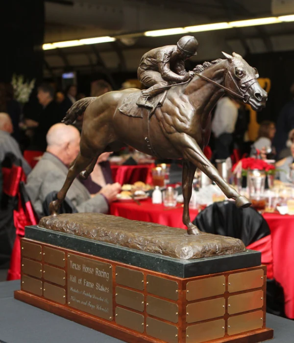 Bronze horse racing trophy with rider on wooden and metal base, displayed on a table at the Texas Horse Racing Annual Gala, with seated attendees in the background.