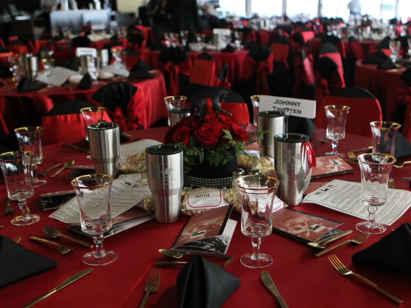 A banquet table at the Texas Horse Racing Annual Gala set with a red tablecloth, black napkins, glassware, silverware, stainless cups, programs, popcorn bags, and a red rose centerpiece; place cards visible.